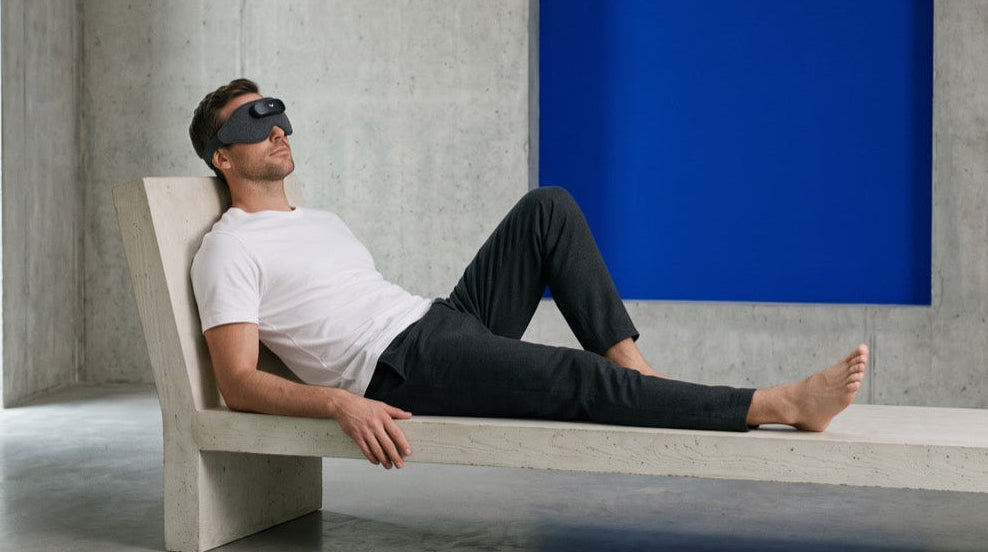 A man wearing a the Domayn Mask reclines on a minimalist concrete daybed in a modern concrete-walled room with a bold blue accent panel.