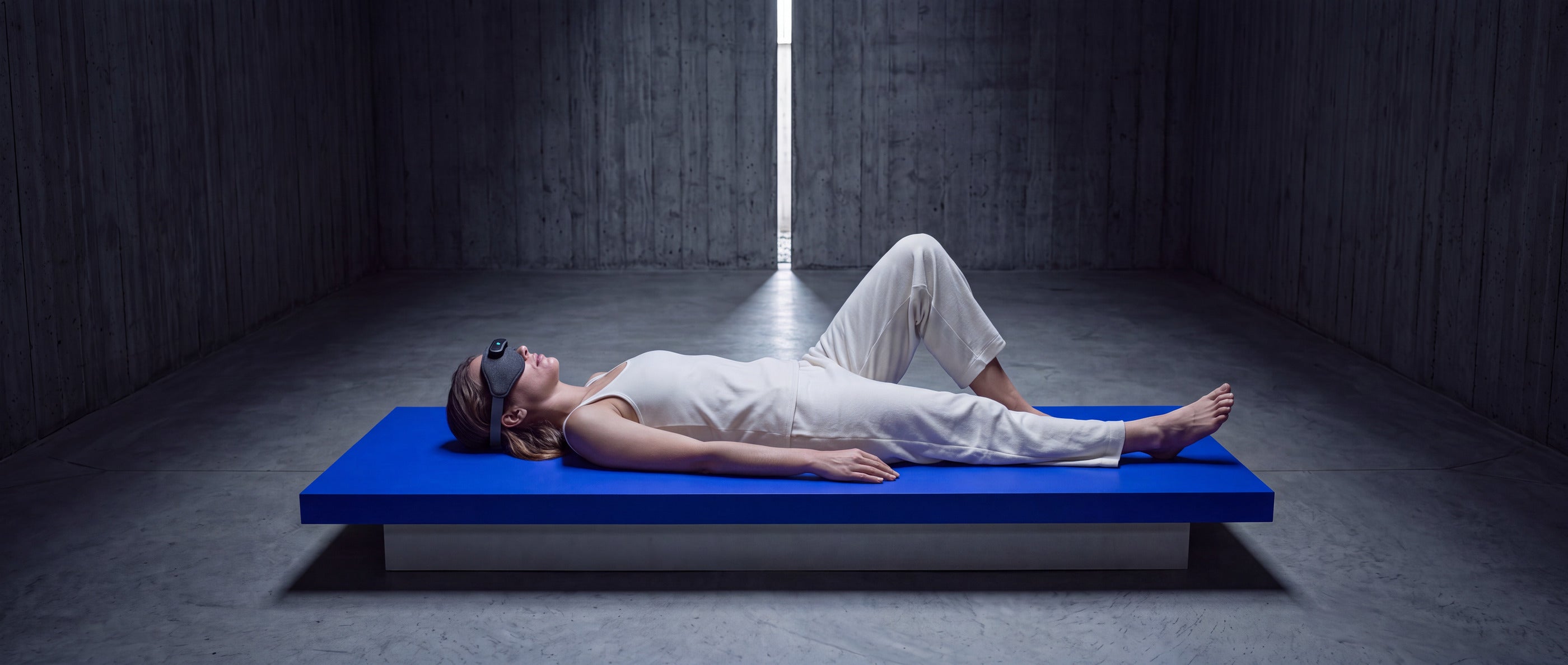 Person lying on a raised platform wearing the Domayn Mask, in a bare concrete room lit by a single vertical window